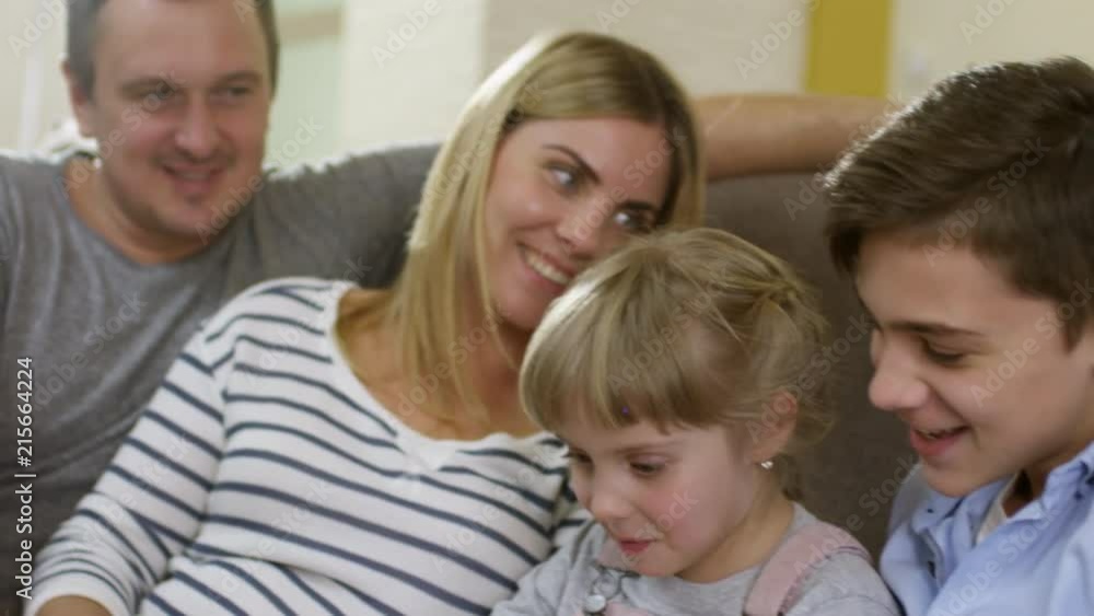 Happy mother, father, little daughter and teenage son sitting together on sofa at home, smiling and playing on digital tablet Happy mother, father, little daughter and teenage son sitting together on sofa at home, smiling and playing on digital tablet