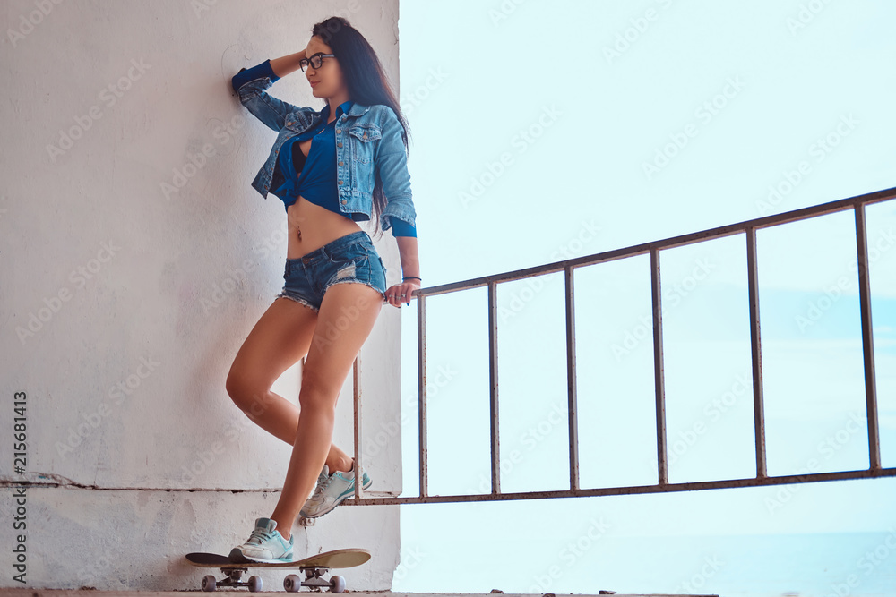 Sexy brunette girl wearing short shorts and jeans jacket standing on a  skateboard and leaning on a guardrail against a sea coast. Stock Photo |  Adobe Stock