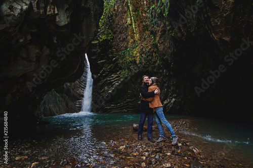 Romantic couple in mountains. Man and woman standing and hugging near waterfall. Active lifestyle in nature.