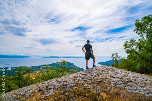 Man watching from the top of a mountain over the water and forest, standing on a rock