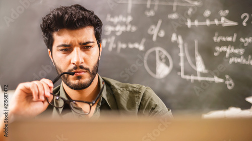 Handsome multicultural bearded businessman in glasses and green shirt sitting behind the laptop and thinking about new project in the office on the some diagrams background.