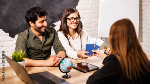 Young cute couple choosing a trip with agent sitting at the travel agency office with world map on the background