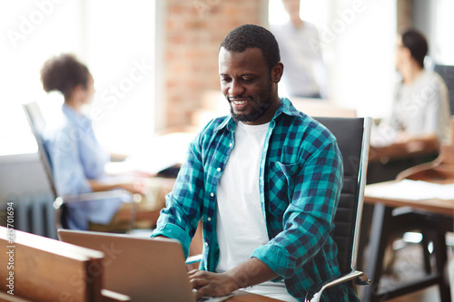 African smiling businessman in casual clothes sitting at his workplace and typing on laptop