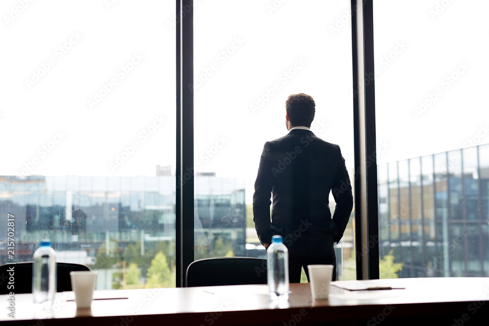 Fototapeta premium Contemplative businessman in formal suit holding hand in pockets and looking out panoramic window while waiting for conference in meeting room