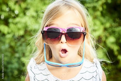 Child cute girl having several sunglasses on her face and doing fun during a hot summer day. Greenery background