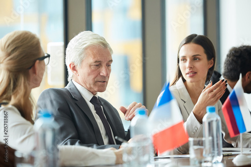 Serious thoughtful handsome senior male politician in suit sitting at conference table with national flags and talking to colleagues while discussing agenda