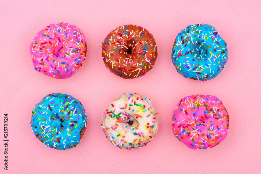 Six assorted donuts with pastel colored icing and sprinkles against a soft pink background. Minimal concept.