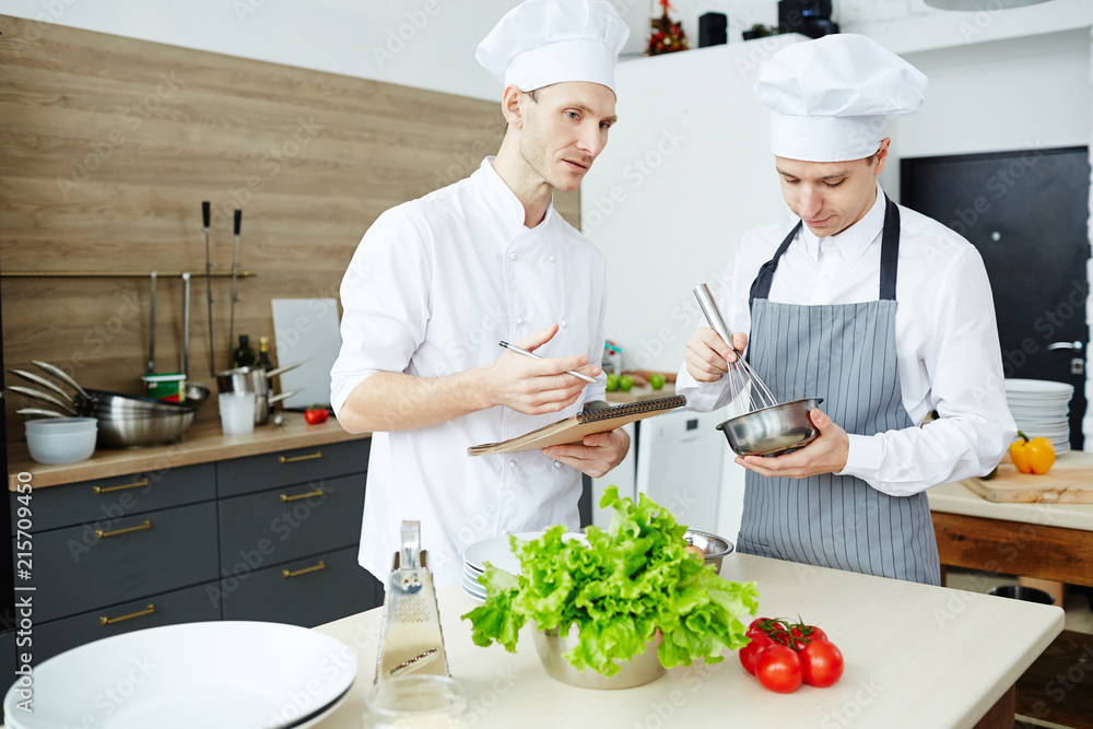 Serious confident handsome young chef explaining how to cook sauce to young man, male intern in apron whipping meal in bowl and listening to teacher