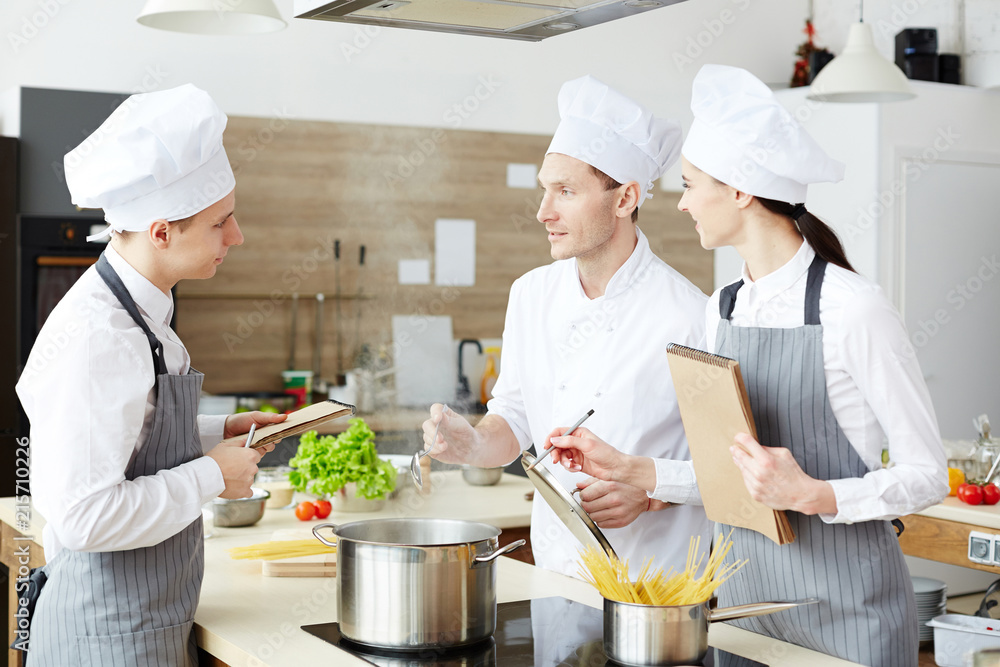 Confident experienced chef in white uniform explaining cooking techniques while showing how to cook tasty pasta, interns asking questions and making notes Confident experienced chef in white uniform explaining cooking techniques while showing how to cook tasty pasta, interns asking questions and making notes