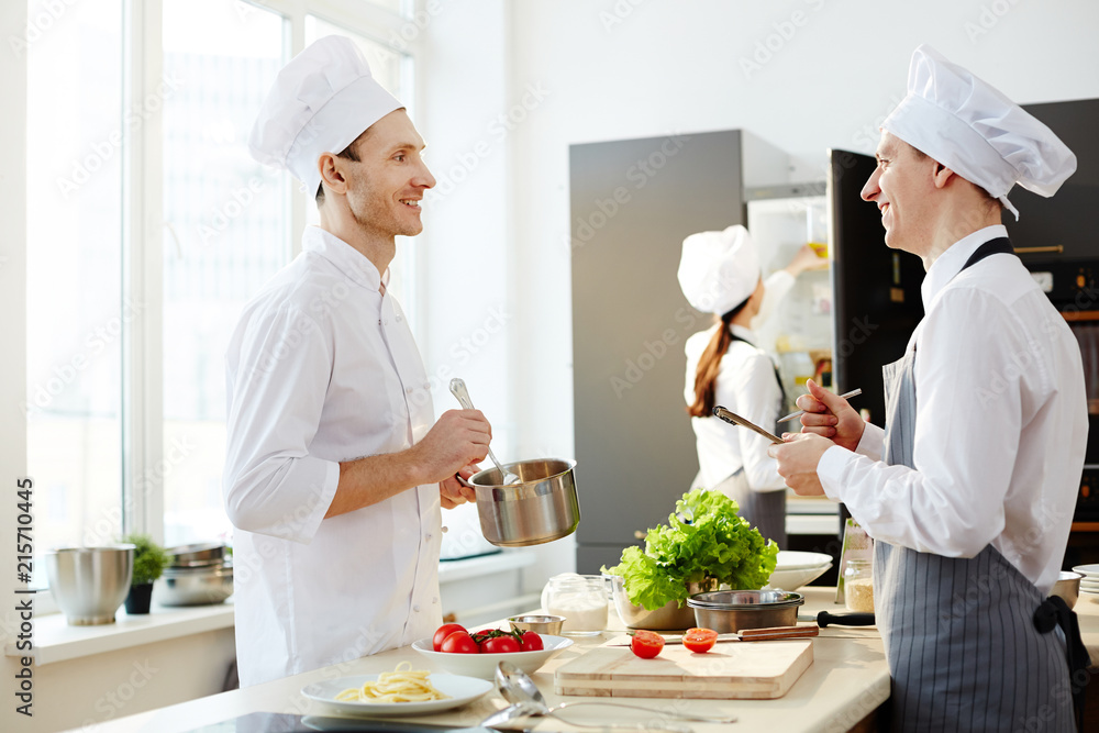 Positive handsome chef in white uniform stirring sauce for pasta in pan while showing how to cook to young intern in apron at commercial kitchen Positive handsome chef in white uniform stirring sauce for pasta in pan while showing how to cook to young intern in apron at commercial kitchen