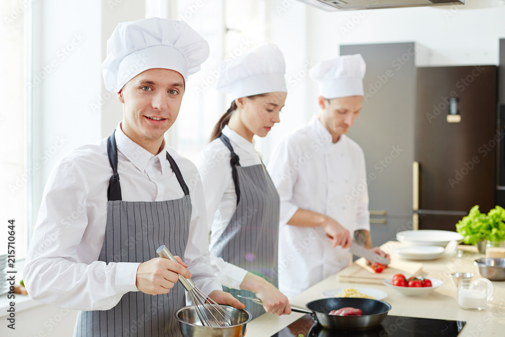 Smiling handsome young cook in apron and chefs hat whipping ingredients in bowl and looking at camera while his colleagues cooking in background Smiling handsome young cook in apron and chefs hat whipping ingredients in bowl and looking at camera while his colleagues cooking in background