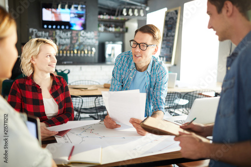 Team of creative business professionals discussing ideas while collaborating on startup project during meeting in modern office, focus on cheerful young man leading group