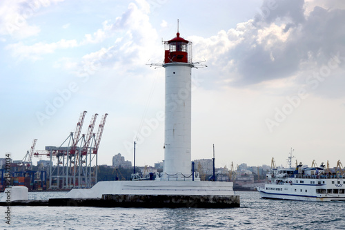 Lighthouse in the sea against the backdrop of a cargo port. Summer seascape with a white lighthouse with red top. Black Sea. Sea Port of Odessa. © Ирина Абраменко