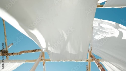 White linen cloth on wooden poles swings in the wind. Against the blue sky and green trees and fields.