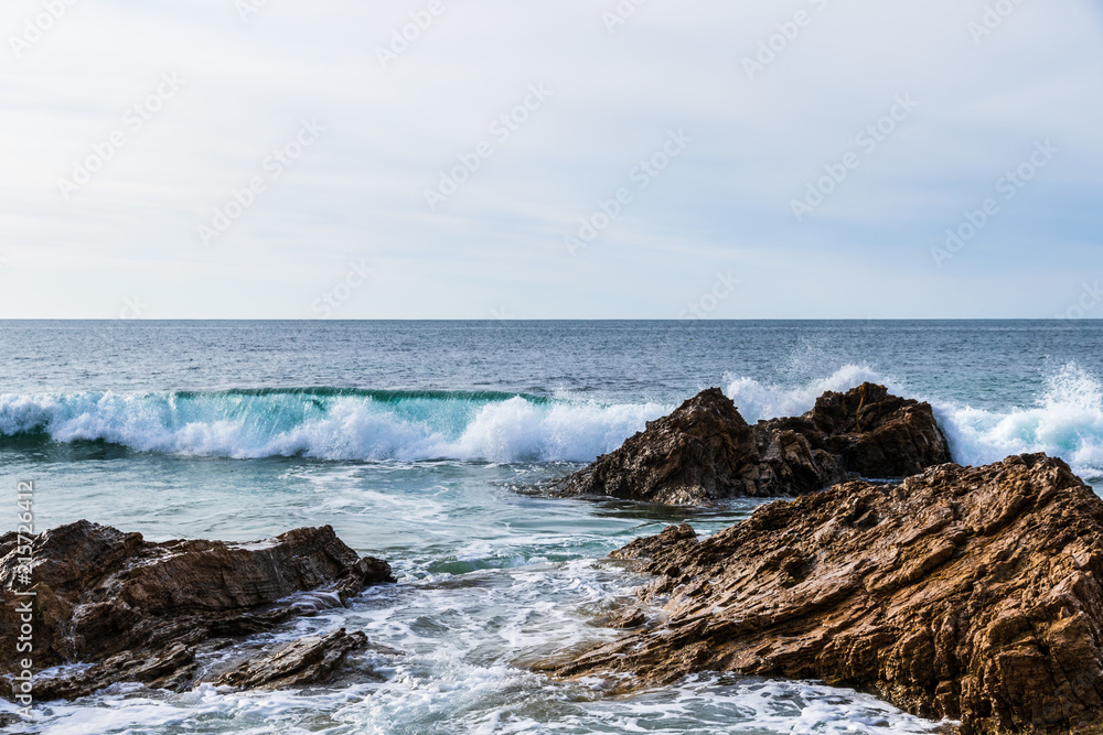 Wave crashing behind rocks near the beach of Crystal Cove State Park in Laguna Beach, California. Foam from an earlier wave is in the foreground; Pacific ocean is in the background.