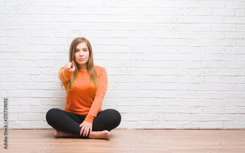Young adult woman sitting on the floor in autumn over white brick wall pointing with finger to the camera and to you, hand sign, positive and confident gesture from the front