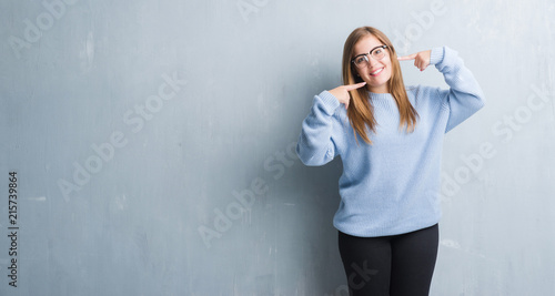 Young adult woman over grey grunge wall wearing glasses smiling confident showing and pointing with fingers teeth and mouth. Health concept.