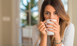 Beautiful young woman drinking coffee while holding cup with hands looking at the camera. Breakfast at home