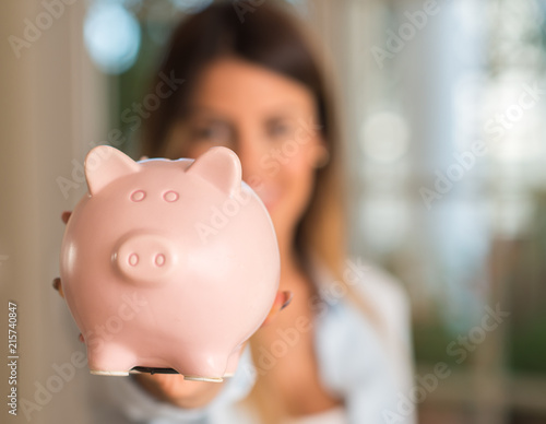 Close up of beautiful young woman holding piggy bank at home, very happy with a smile because of profit. Business concept.