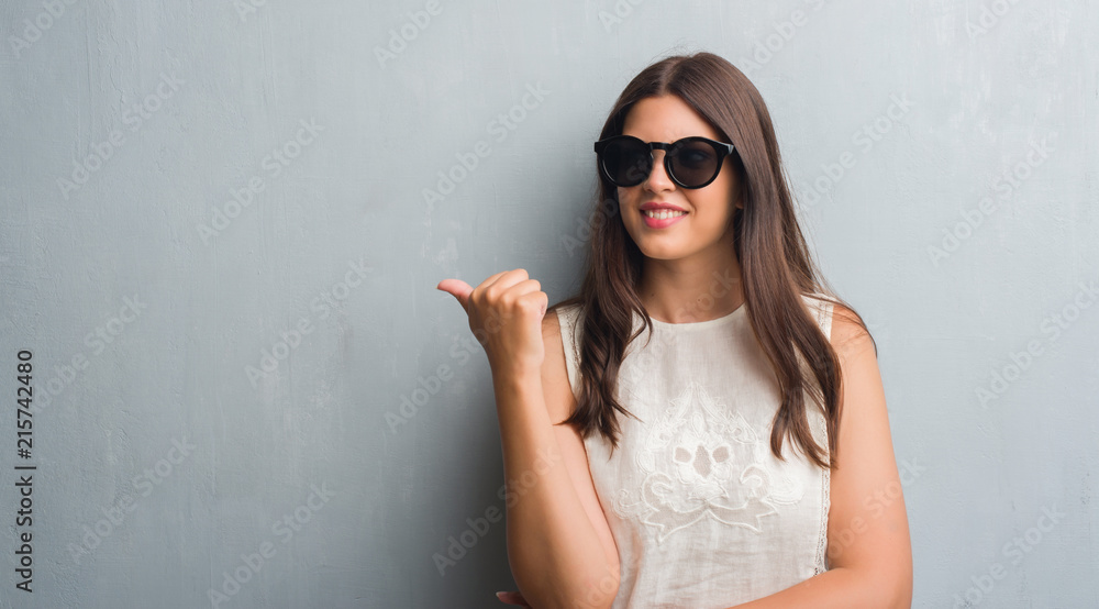 Young brunette woman over grunge grey wall wearing fashion sunglasses pointing and showing with thumb up to the side with happy face smiling Young brunette woman over grunge grey wall wearing fashion sunglasses pointing and showing with thumb up to the side with happy face smiling