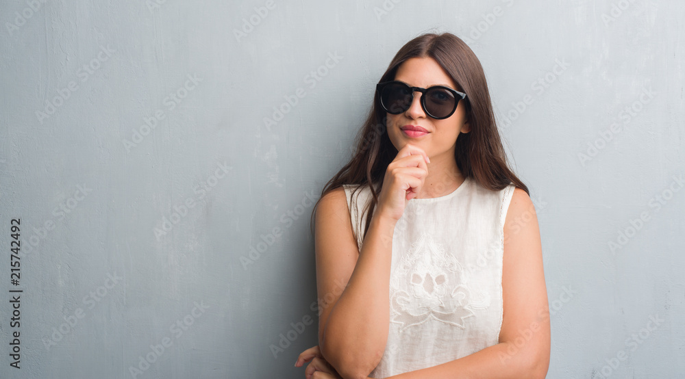 Young brunette woman over grunge grey wall wearing fashion sunglasses serious face thinking about question, very confused idea Young brunette woman over grunge grey wall wearing fashion sunglasses serious face thinking about question, very confused idea