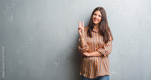 Young brunette woman over grunge grey wall showing and pointing up with fingers number three while smiling confident and happy. © Krakenimages.com