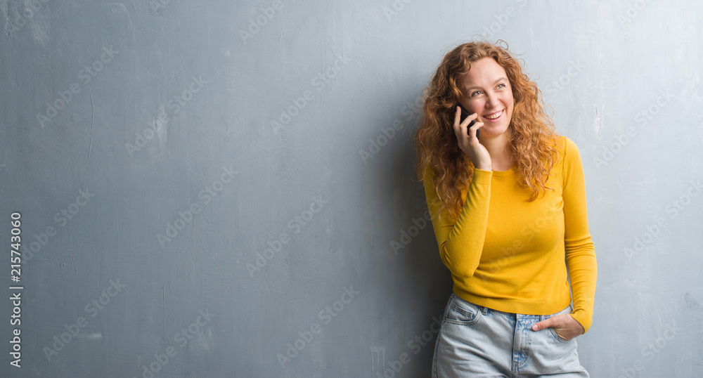 Young redhead woman over grey grunge wall talking on the phone with a happy face standing and smiling with a confident smile showing teeth Young redhead woman over grey grunge wall talking on the phone with a happy face standing and smiling with a confident smile showing teeth