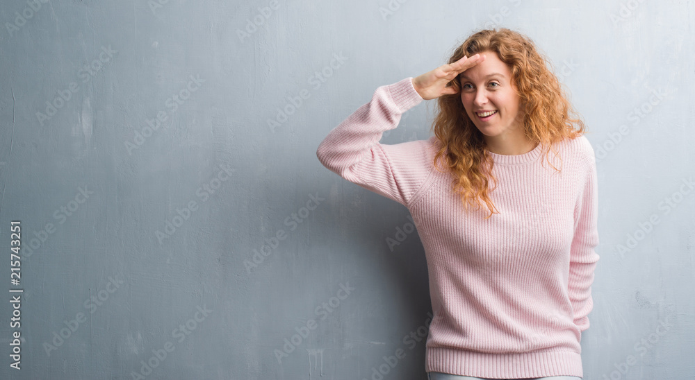 Young redhead woman over grey grunge wall wearing pink sweater very happy and smiling looking far away with hand over head. Searching concept.