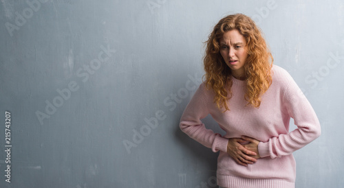 Young redhead woman over grey grunge wall wearing pink sweater with hand on stomach because indigestion, painful illness feeling unwell. Ache concept.