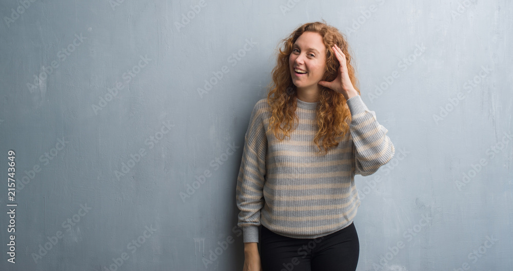 Fototapeta premium Young redhead woman over grey grunge wall smiling with hand over ear listening an hearing to rumor or gossip. Deafness concept.