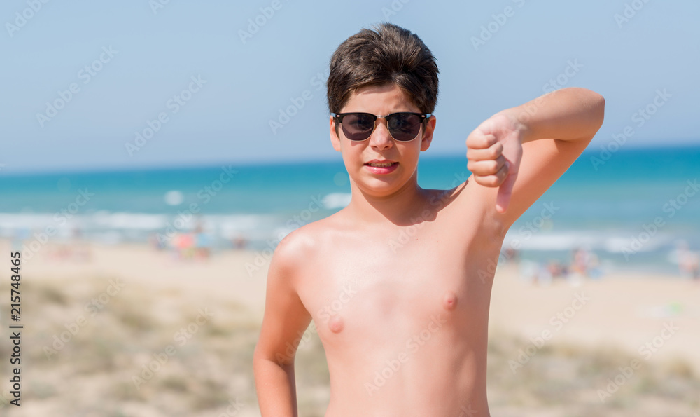 Young child on holidays at the beach with angry face, negative sign showing dislike with thumbs down, rejection concept Young child on holidays at the beach with angry face, negative sign showing dislike with thumbs down, rejection concept