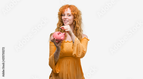 Young redhead woman holding piggy bank with a happy face standing and smiling with a confident smile showing teeth