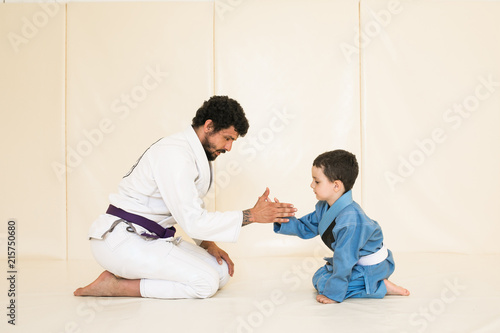 Father and little kid son are engaged in wrestling jiu-jitsu in the gym in a kimono. Trainer teaches child the methods and positions of single combat, karate or aikido. Sport and healthy in family