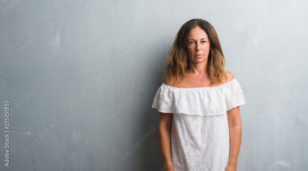 Middle age hispanic woman standing over grey grunge wall with serious expression on face. Simple and natural looking at the camera. Middle age hispanic woman standing over grey grunge wall with serious expression on face. Simple and natural looking at the camera.