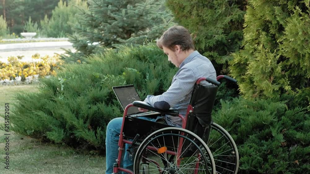 A disabled man is sitting in a wheelchair and working on a laptop in the park A disabled man is sitting in a wheelchair and working on a laptop in the park