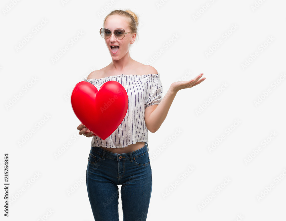 Blonde teenager woman holding red heart very happy and excited, winner expression celebrating victory screaming with big smile and raised hands Blonde teenager woman holding red heart very happy and excited, winner expression celebrating victory screaming with big smile and raised hands