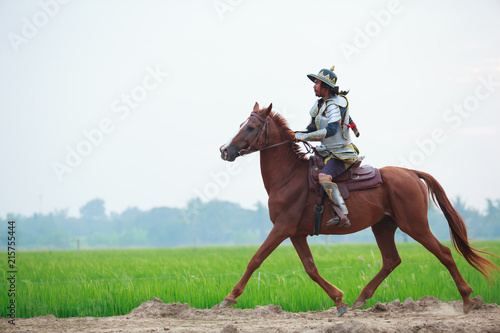 Asian Thai Warrior in traditional armor suit riding horse in rural farm background. Vintage Retro war costume concept.