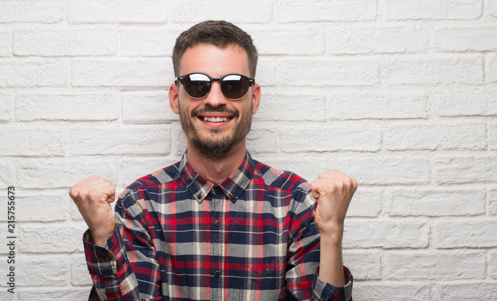 Young adult man wearing sunglasses standing over white brick wall screaming proud and celebrating victory and success very excited, cheering emotion Young adult man wearing sunglasses standing over white brick wall screaming proud and celebrating victory and success very excited, cheering emotion