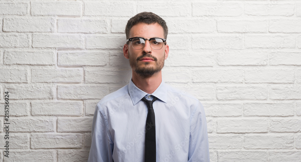 Young adult business man standing over white brick wall with a confident expression on smart face thinking serious Young adult business man standing over white brick wall with a confident expression on smart face thinking serious