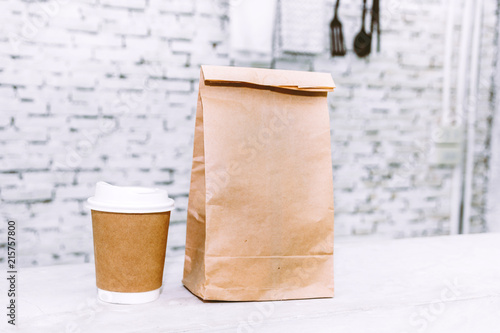 Cup of coffee and blank paper bag with copy space for your brand on table against white brick wall background