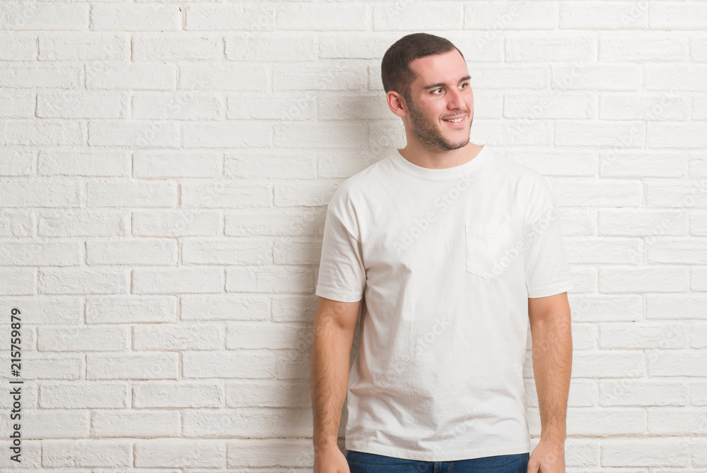 Young caucasian man standing over white brick wall looking away to side with smile on face, natural expression. Laughing confident. Young caucasian man standing over white brick wall looking away to side with smile on face, natural expression. Laughing confident.