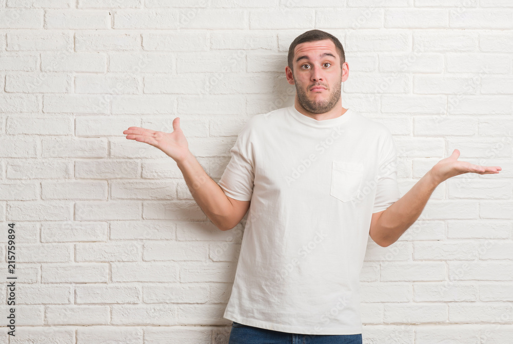 Young caucasian man standing over white brick wall clueless and confused expression with arms and hands raised. Doubt concept. Young caucasian man standing over white brick wall clueless and confused expression with arms and hands raised. Doubt concept.
