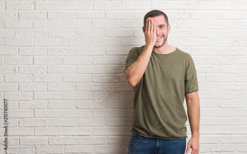 Young caucasian man standing over white brick wall covering one eye with hand with confident smile on face and surprise emotion.