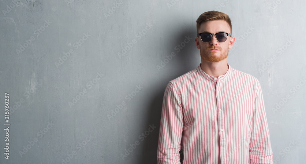 Obraz premium Young redhead man over grey grunge wall wearing sunglasses with a confident expression on smart face thinking serious