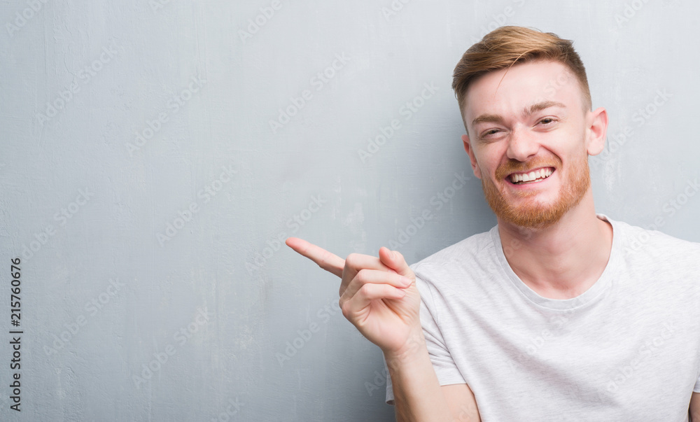 Young redhead man over grey grunge wall very happy pointing with hand and finger to the side Young redhead man over grey grunge wall very happy pointing with hand and finger to the side