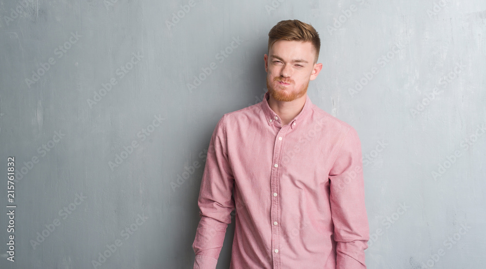 Young redhead man over grey grunge wall wearing pink shirt puffing cheeks with funny face. Mouth inflated with air, crazy expression.