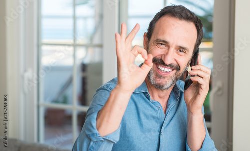 Middle age man using smartphone doing ok sign with fingers, excellent symbol