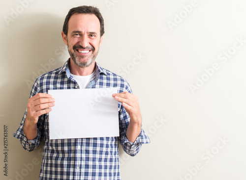 Senior man holding blank paper sheet with a happy face standing and smiling with a confident smile showing teeth photo