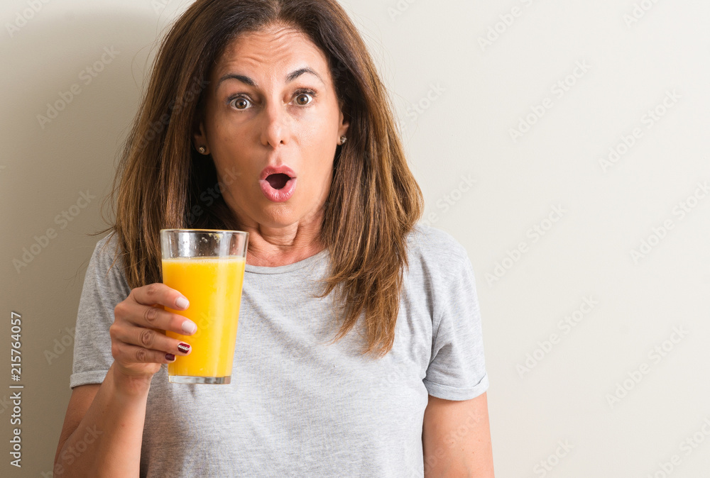 Middle age woman drinking orange juice in a glass scared in shock with a surprise face, afraid and excited with fear expression Middle age woman drinking orange juice in a glass scared in shock with a surprise face, afraid and excited with fear expression