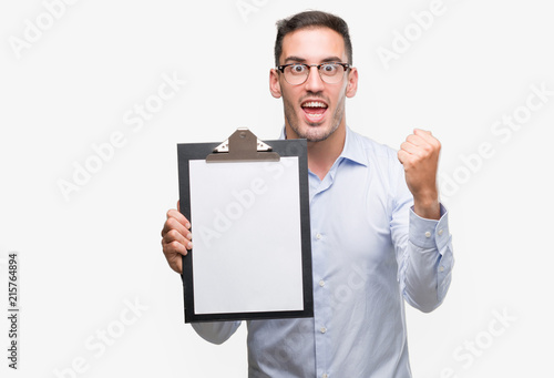 Handsome young business man showing a clipboard screaming proud and celebrating victory and success very excited, cheering emotion